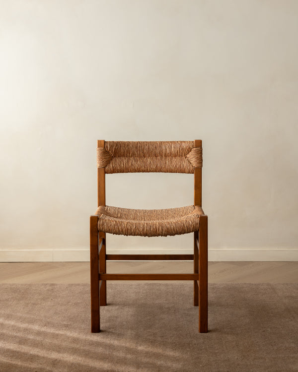 A mid-century modern wooden dining chair with a woven natural fiber seat and backrest, positioned against a minimalist beige wall and textured carpet.