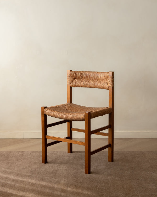 A wooden dining chair with a woven natural-fiber seat and backrest, positioned on a textured beige rug against a neutral beige wall.