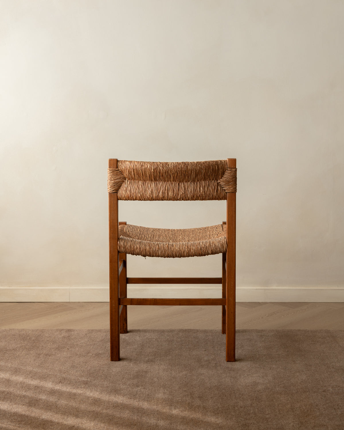 A wooden dining chair with a woven natural-fiber seat and back, positioned against a neutral beige wall and textured brown carpet.