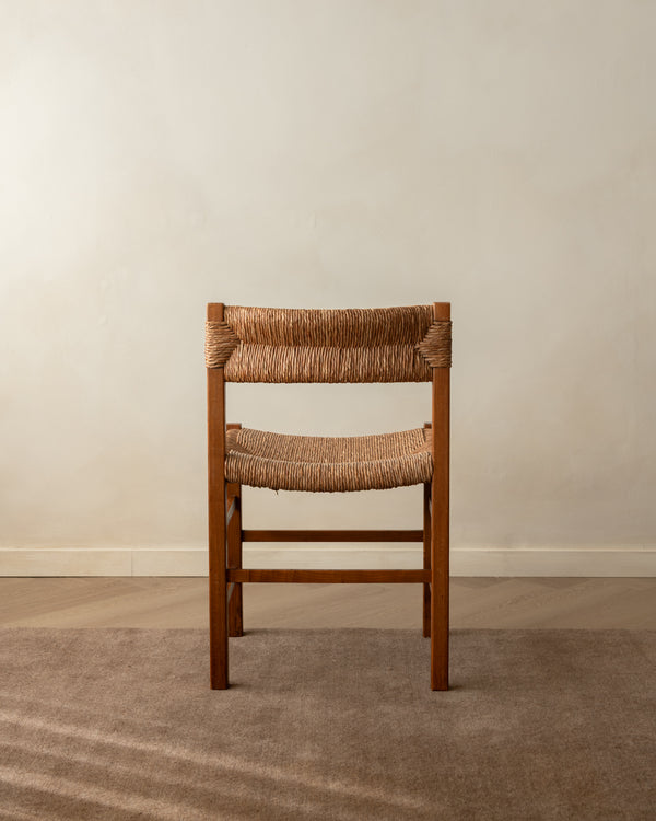 A wooden dining chair with a woven natural-fiber seat and back, positioned against a neutral beige wall and textured brown carpet.