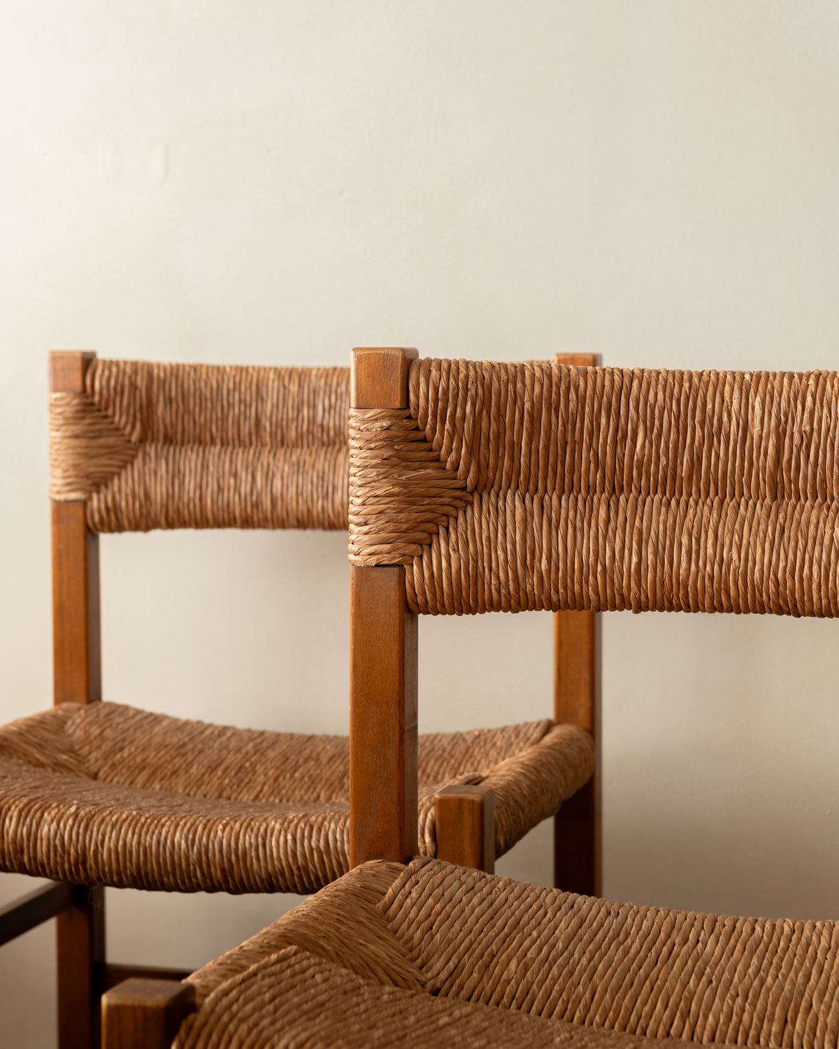 Close-up of two wooden chairs with woven rope seats and backs, shown against a plain light beige wall.