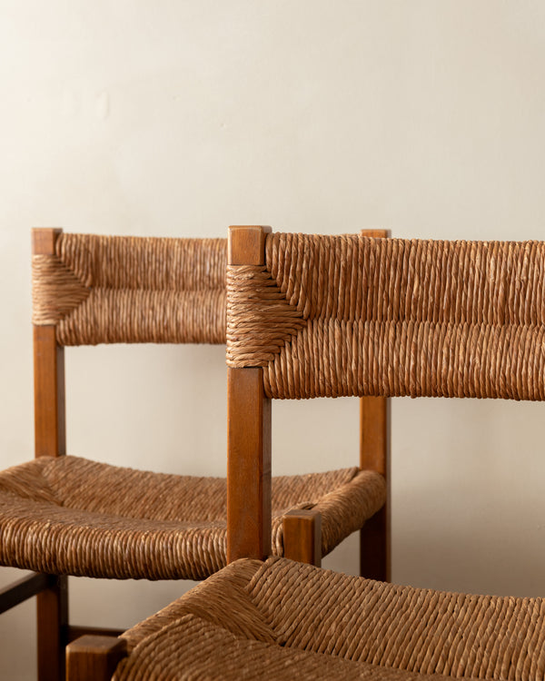 Close-up of two wooden chairs with woven rope seats and backs, shown against a plain light beige wall.