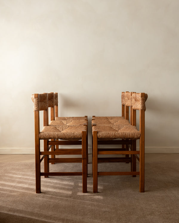 Two wooden dining chairs with woven straw seats and backrests, positioned side-by-side against a plain beige wall and carpeted floor.