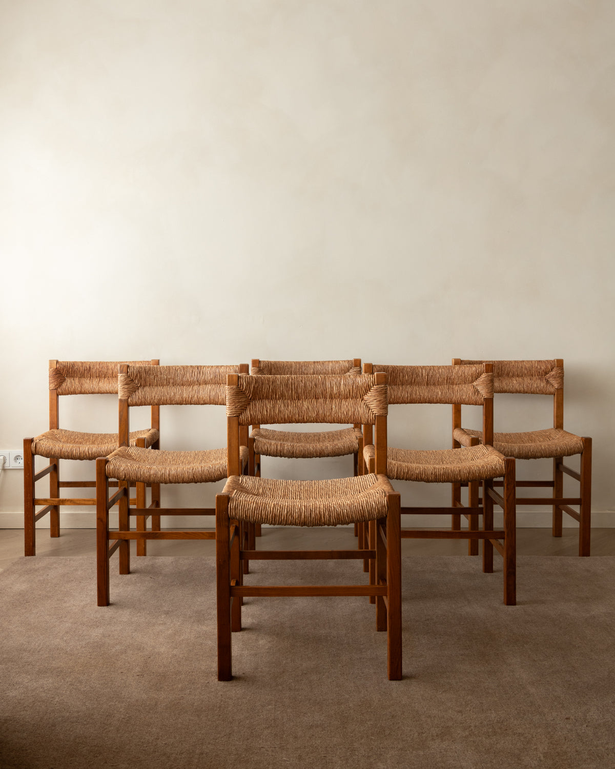 Six wooden dining chairs with woven natural-fiber seats and backrests arranged in two rows against a plain beige wall and carpeted floor.