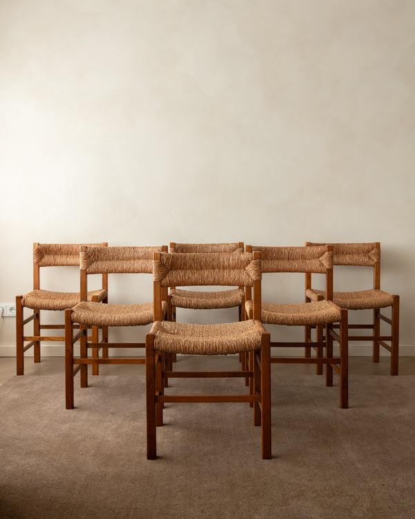 Six wooden dining chairs with woven natural-fiber seats and backrests arranged in two rows against a plain beige wall and carpeted floor.