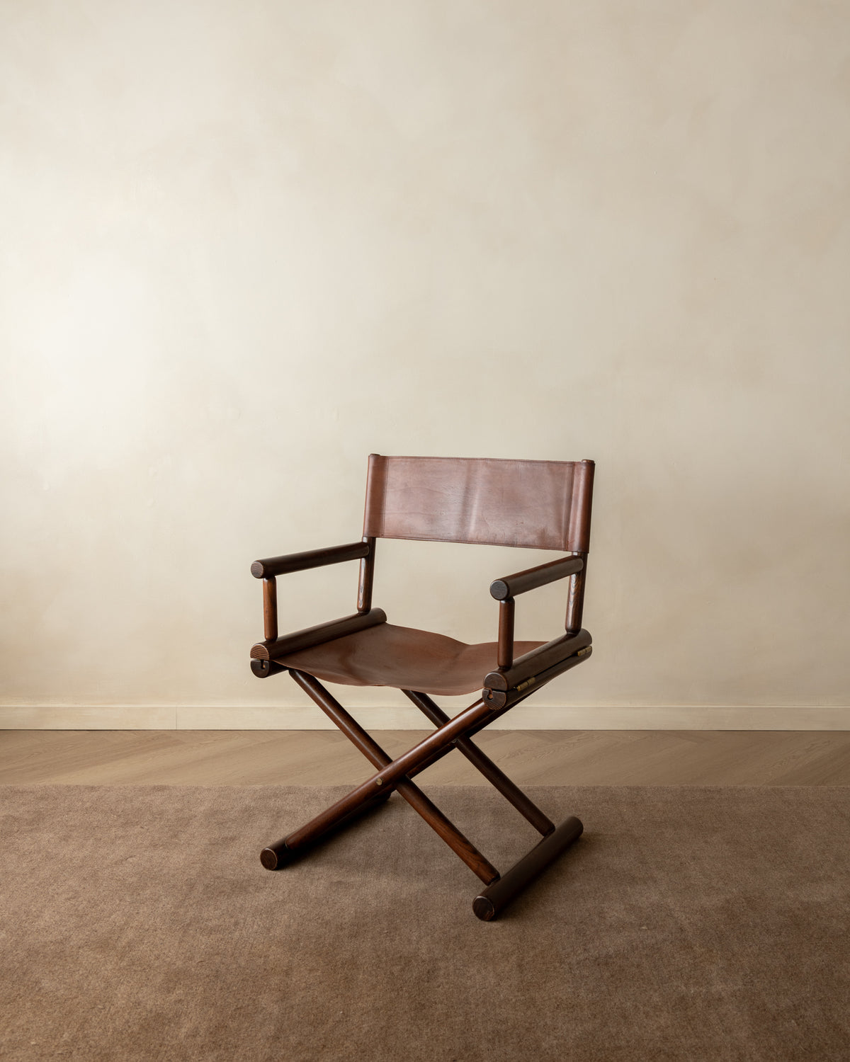 A mid-century modern folding director chair with a dark brown wooden frame and brown leather seat and backrest, set against a neutral beige wall and carpeted floor.