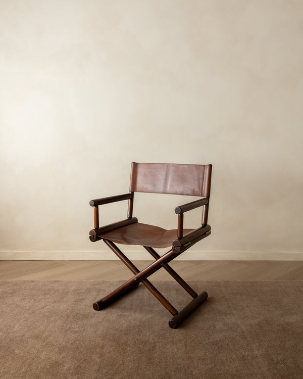A mid-century modern folding director chair with a dark brown wooden frame and brown leather seat and backrest, set against a neutral beige wall and carpeted floor.