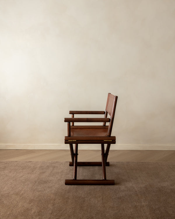 A mid-century modern wooden chair with a dark brown frame and a reddish-brown upholstered seat and back, positioned against a minimalist beige wall and carpeted floor.