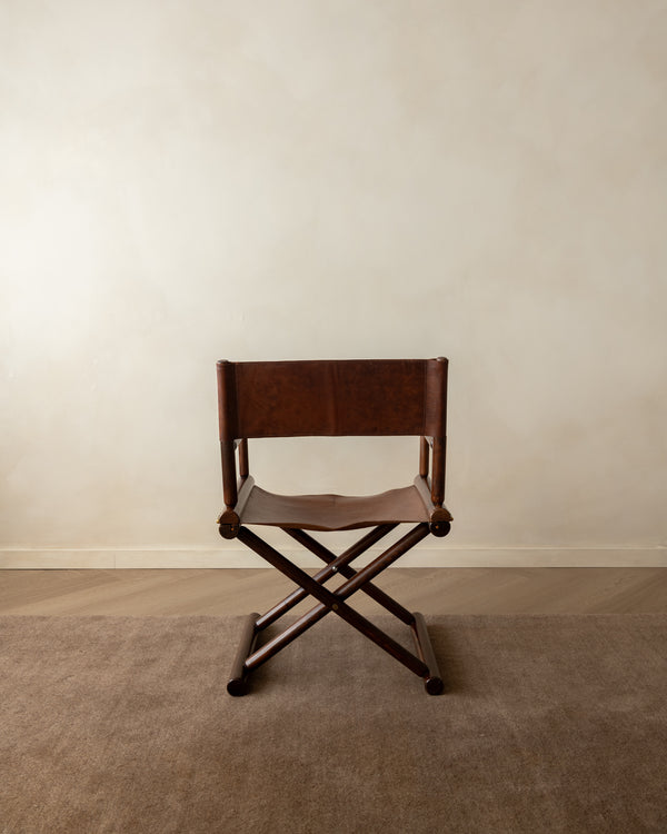 A dark brown leather and wood folding director chair with a slatted back, positioned on a textured beige carpet against a neutral beige wall.