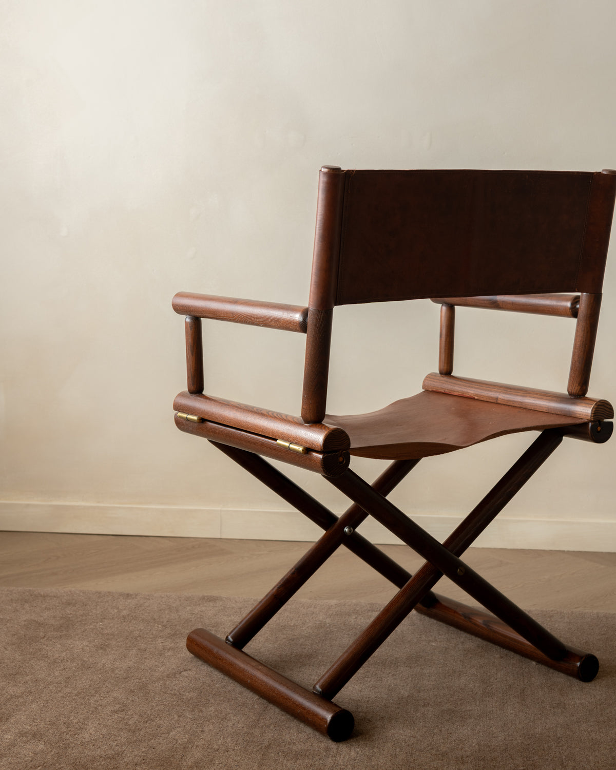 A mid-century modern wooden folding director chair with a dark brown leather seat and backrest, positioned on a textured beige carpet against a neutral beige wall.