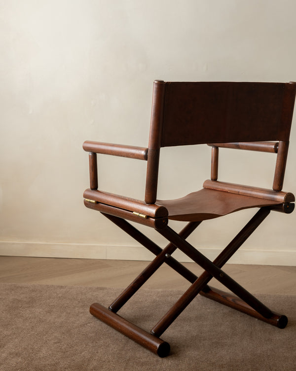 A mid-century modern wooden folding director chair with a dark brown leather seat and backrest, positioned on a textured beige carpet against a neutral beige wall.