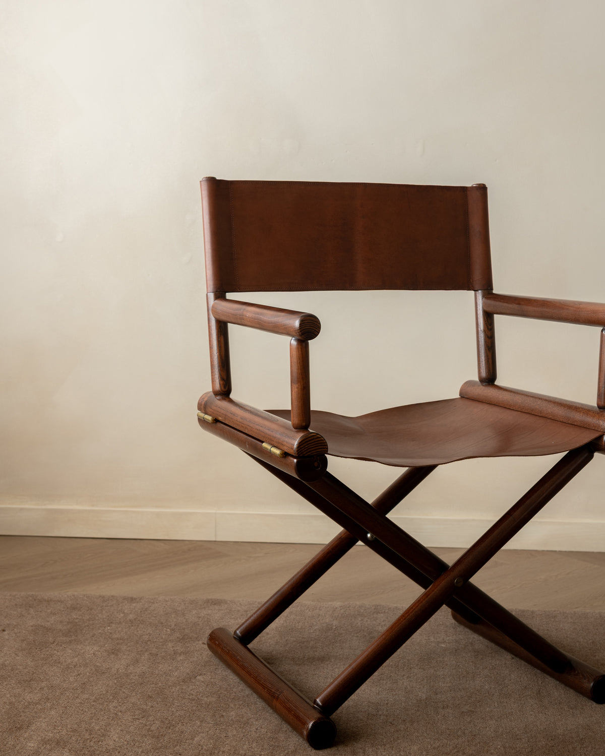 A mid-century modern director's chair with a dark brown wooden frame and brown leather seat and backrest, set against a neutral beige wall and carpeted floor.