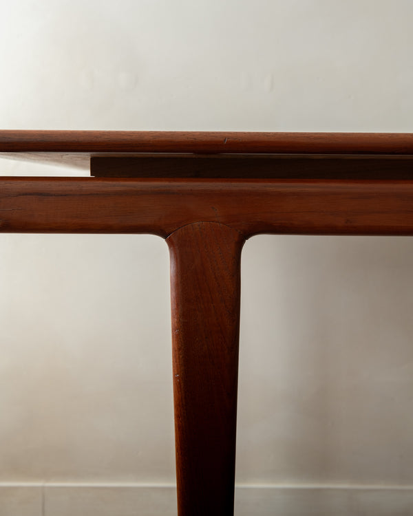 A close-up detail shot of a mid-century modern wooden table, showing the rich reddish-brown grain of the teak wood on the leg and apron, with a light-colored tabletop surface.