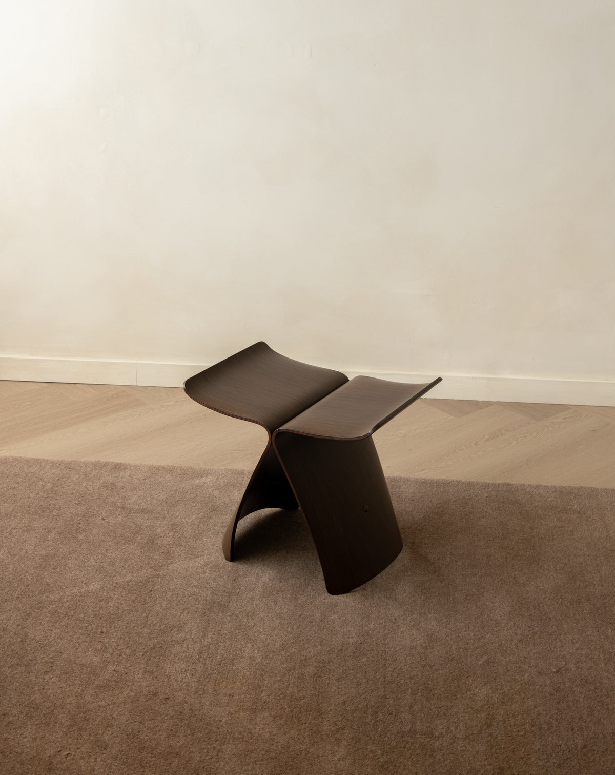 A sculptural dark brown wooden stool with a curved, wave-like design, set on a brown textured rug against a neutral beige wall and light wood floor.