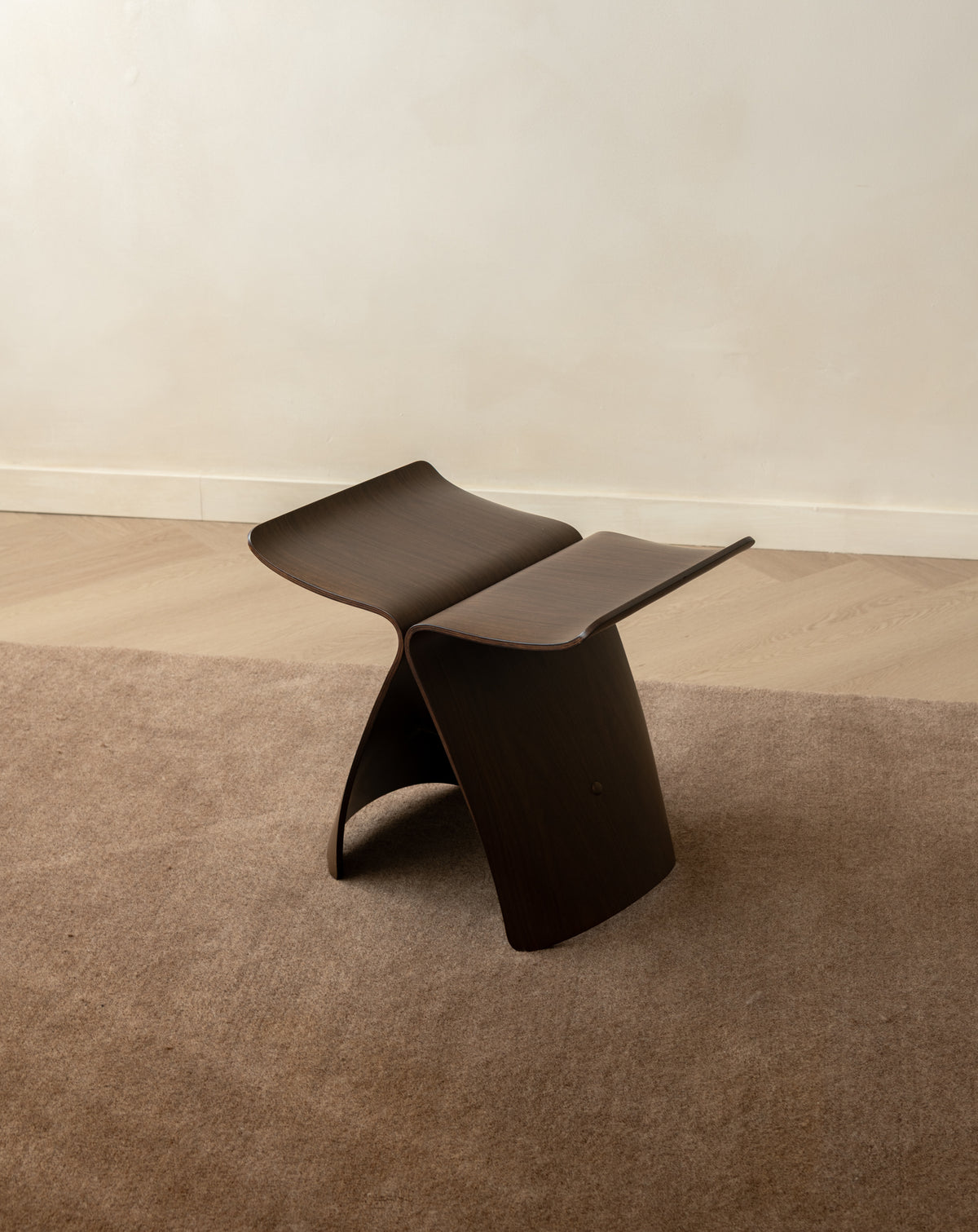 A dark brown sculptural folding stool with a curved, wave-like design, positioned on a textured brown rug against a neutral beige wall and light wood floor.