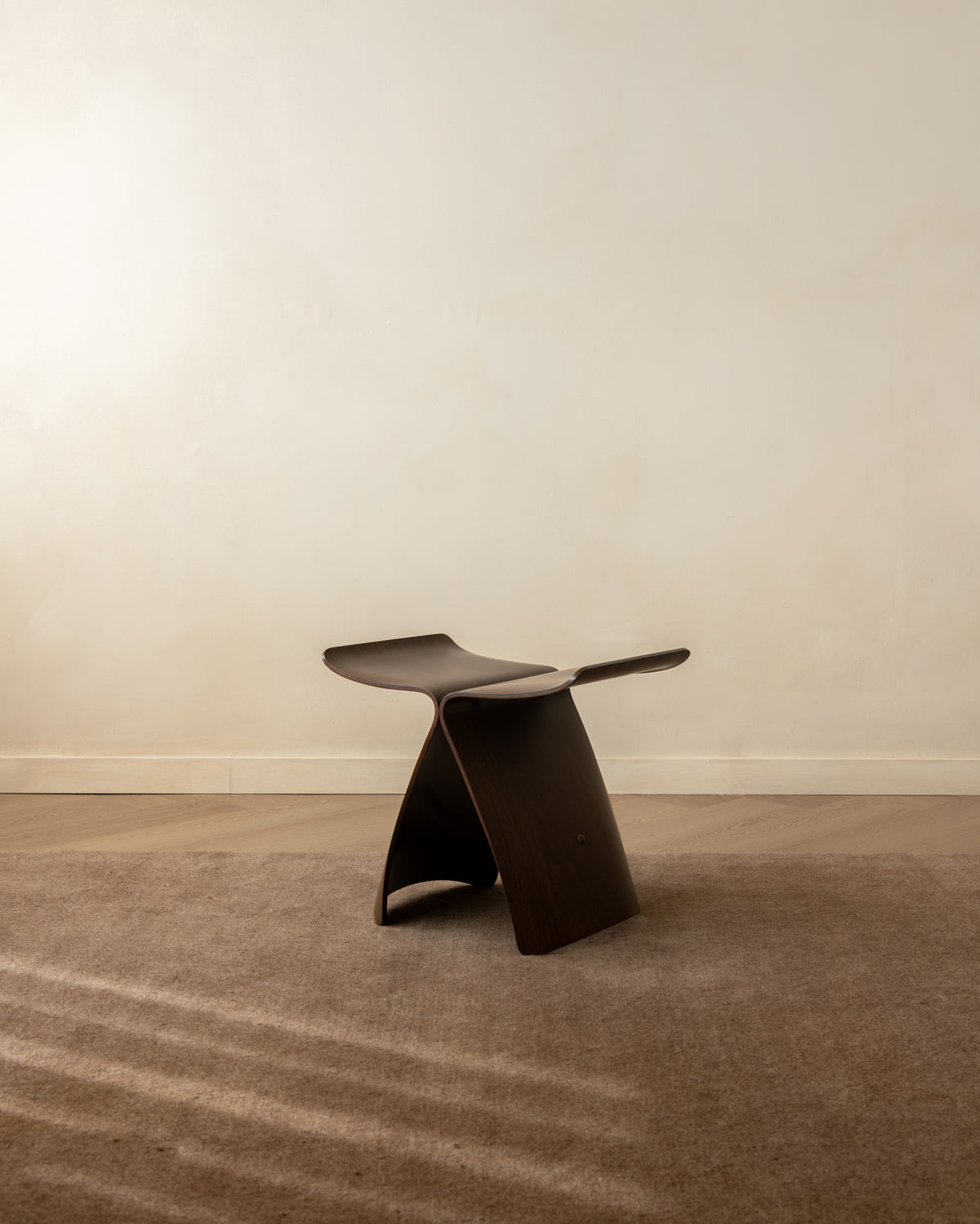 A dark brown sculptural wooden stool with a curved seat and integrated side table extension, set on a textured brown carpet against a plain beige wall.