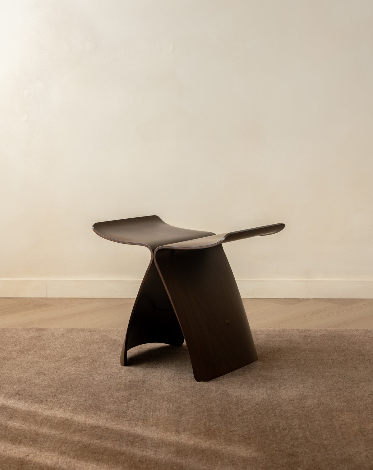 A sculptural dark brown wooden stool with a curved, integrated seat and backrest, set on a textured beige rug against a neutral cream wall.