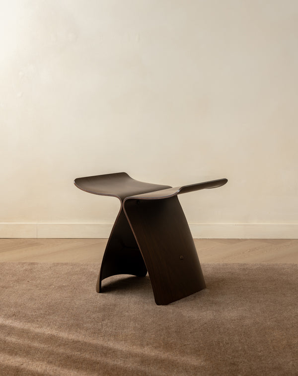 A sculptural dark brown wooden stool with a curved, integrated seat and backrest, set on a textured beige rug against a neutral cream wall.