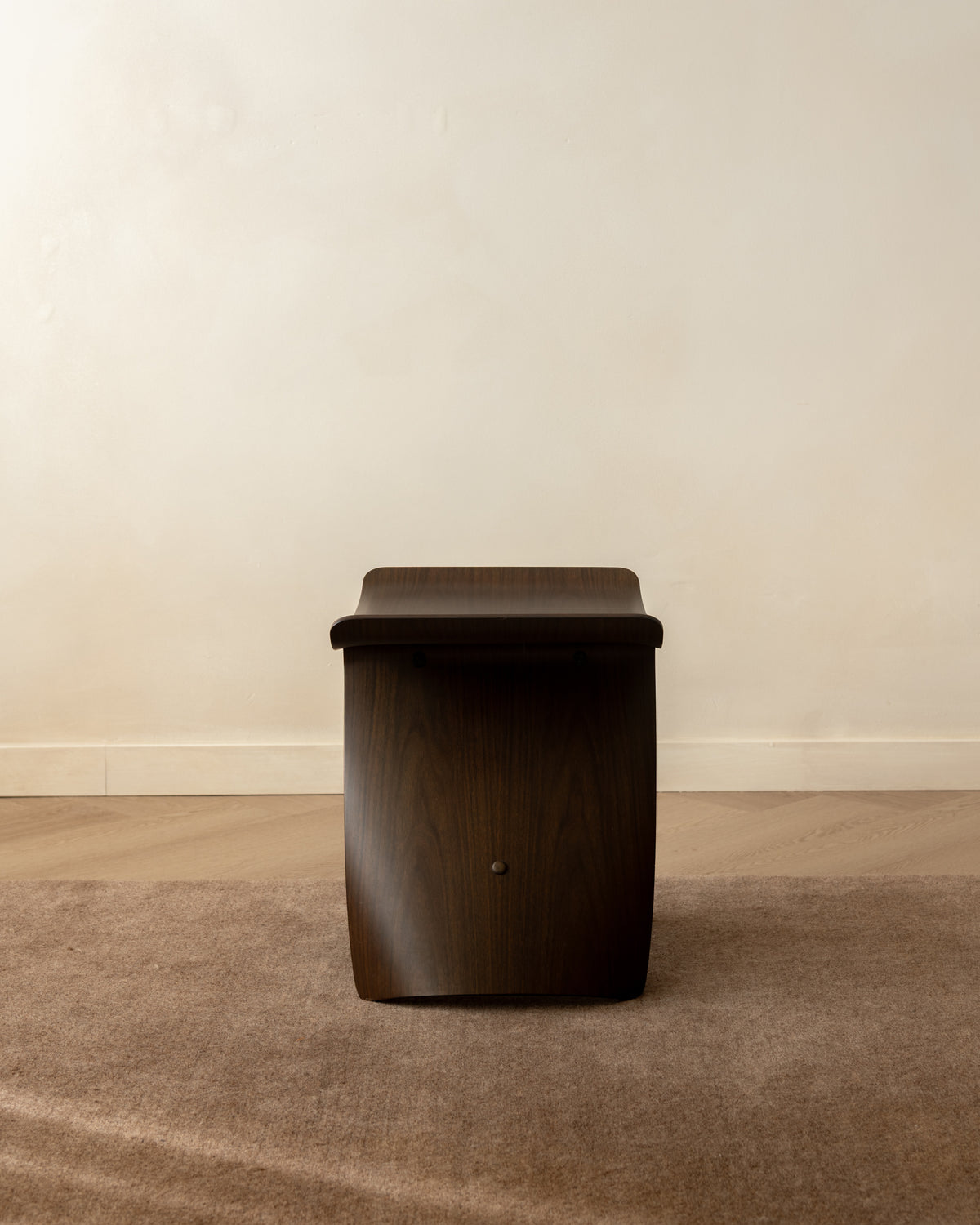 A dark brown wooden bench with a curved, sculptural backrest, positioned on a textured brown rug against a plain beige wall.