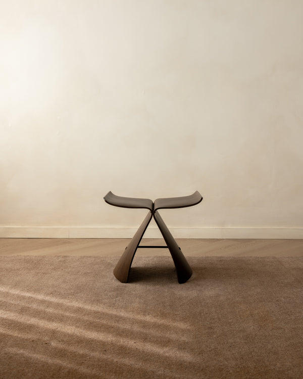 A modern sculptural stool with a dark brown curved seat and sculptural legs, set on a textured beige carpet against a neutral beige wall.