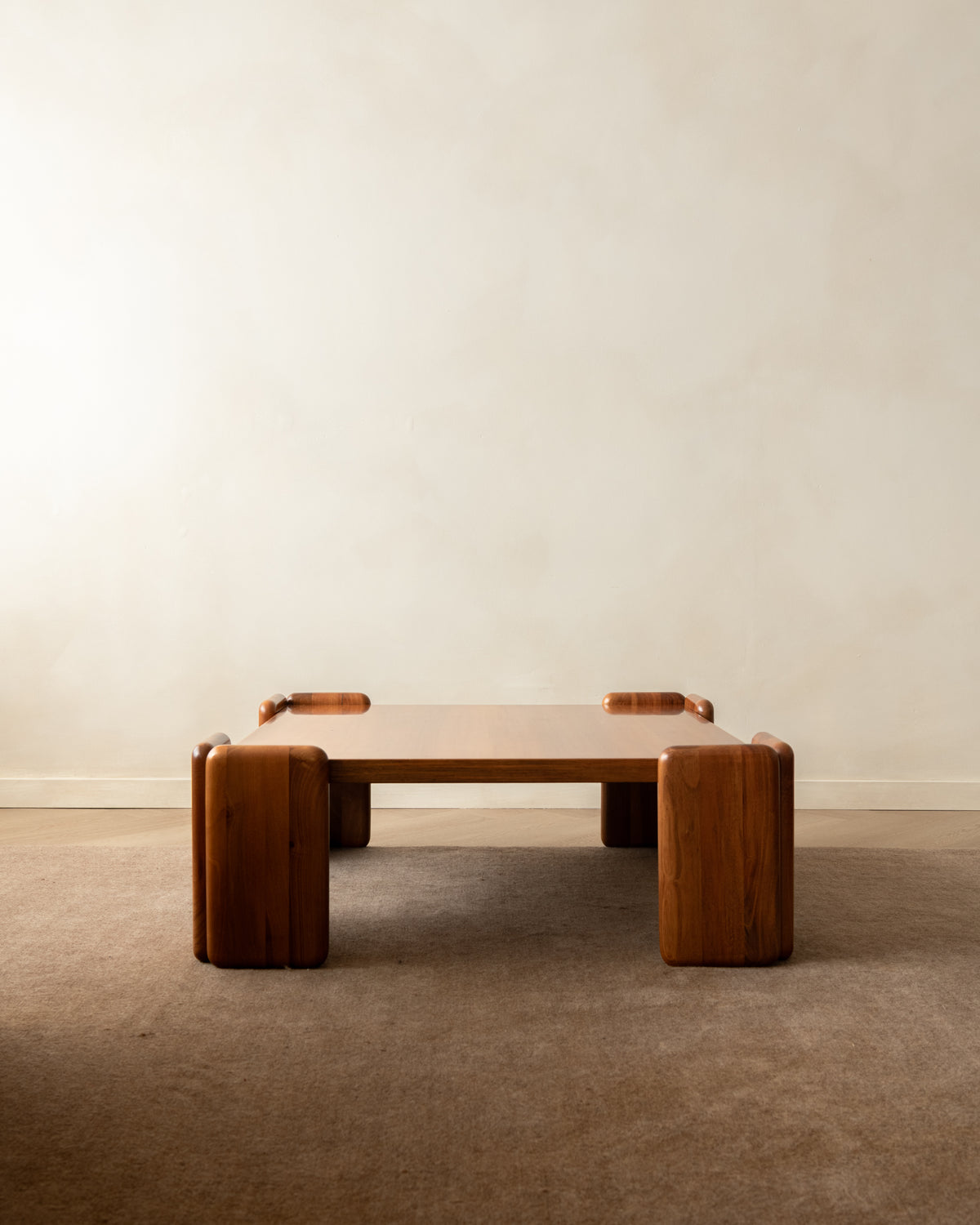 A low wooden coffee table with thick blocky legs and small rectangular side accents, photographed in a minimalist room with a warm beige wall and carpet.