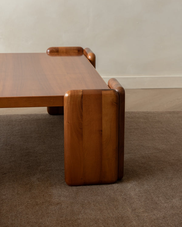 A mid-century modern wooden coffee table with rounded blocky legs and a thick rectangular top, shown on a textured brown carpet against a neutral beige wall.