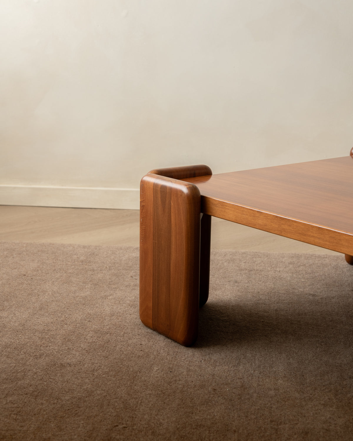 A close-up of a mid-century modern wooden table leg with a rounded rectangular block base and a matching wood tabletop, set on a brown carpet against a beige wall.