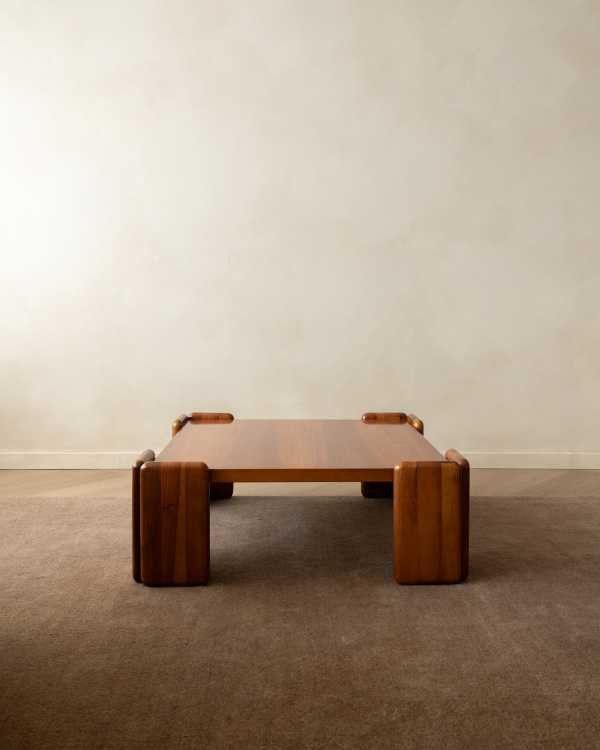 A low rectangular wooden coffee table with thick blocky legs and integrated side handles, shown in a minimalist room with a textured beige wall and carpeted floor.