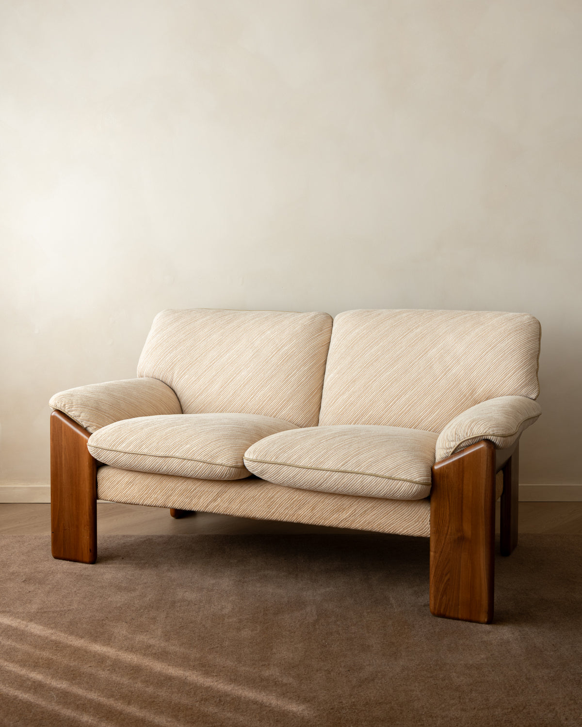 A two-seat upholstered loveseat with cream-colored textured fabric and a medium brown wooden frame, set against a neutral beige wall and brown carpet.