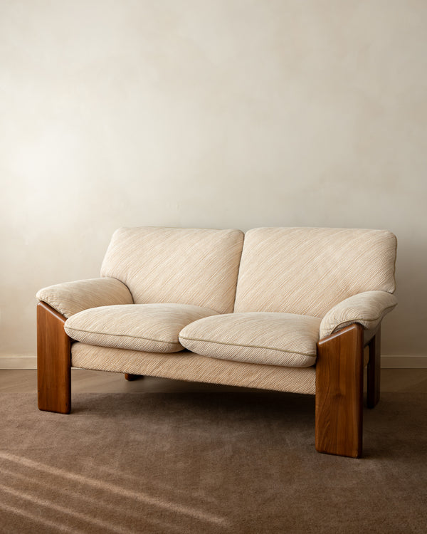 A two-seat upholstered loveseat with cream-colored textured fabric and a medium brown wooden frame, set against a neutral beige wall and brown carpet.