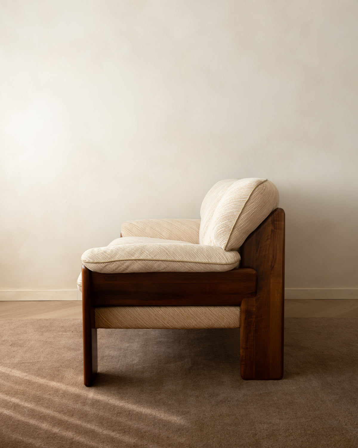 A mid-century modern wooden armchair with cream-colored textured cushions, set against a minimalist beige wall and carpeted floor.
