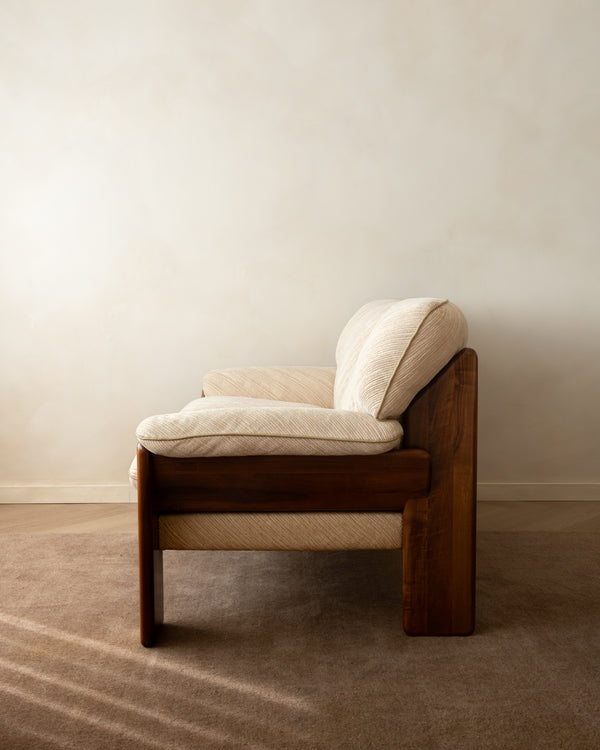 A mid-century modern wooden armchair with cream-colored textured cushions, set against a minimalist beige wall and carpeted floor.
