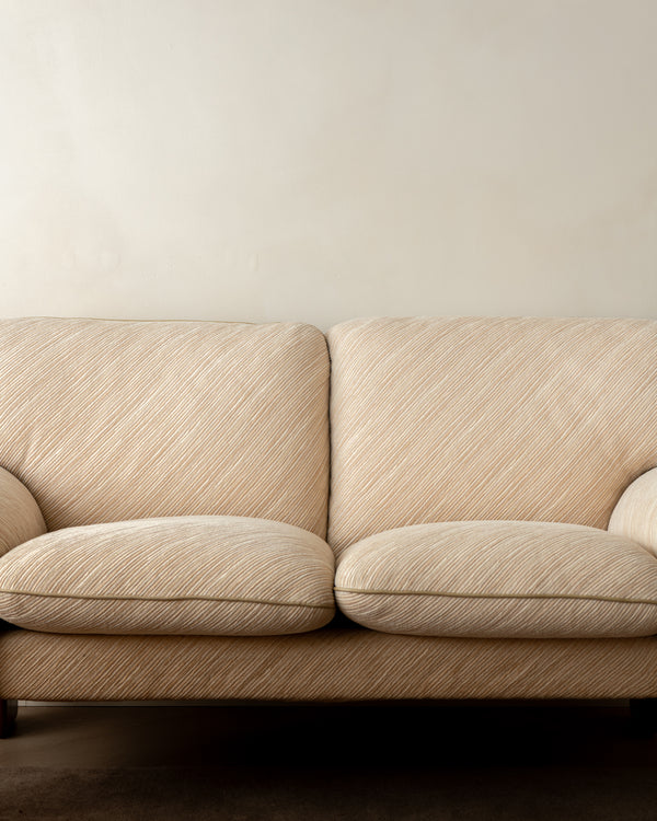 A close-up of a two-seater sofa upholstered in a textured beige fabric with a subtle diagonal ribbed pattern, set against a plain off-white wall.