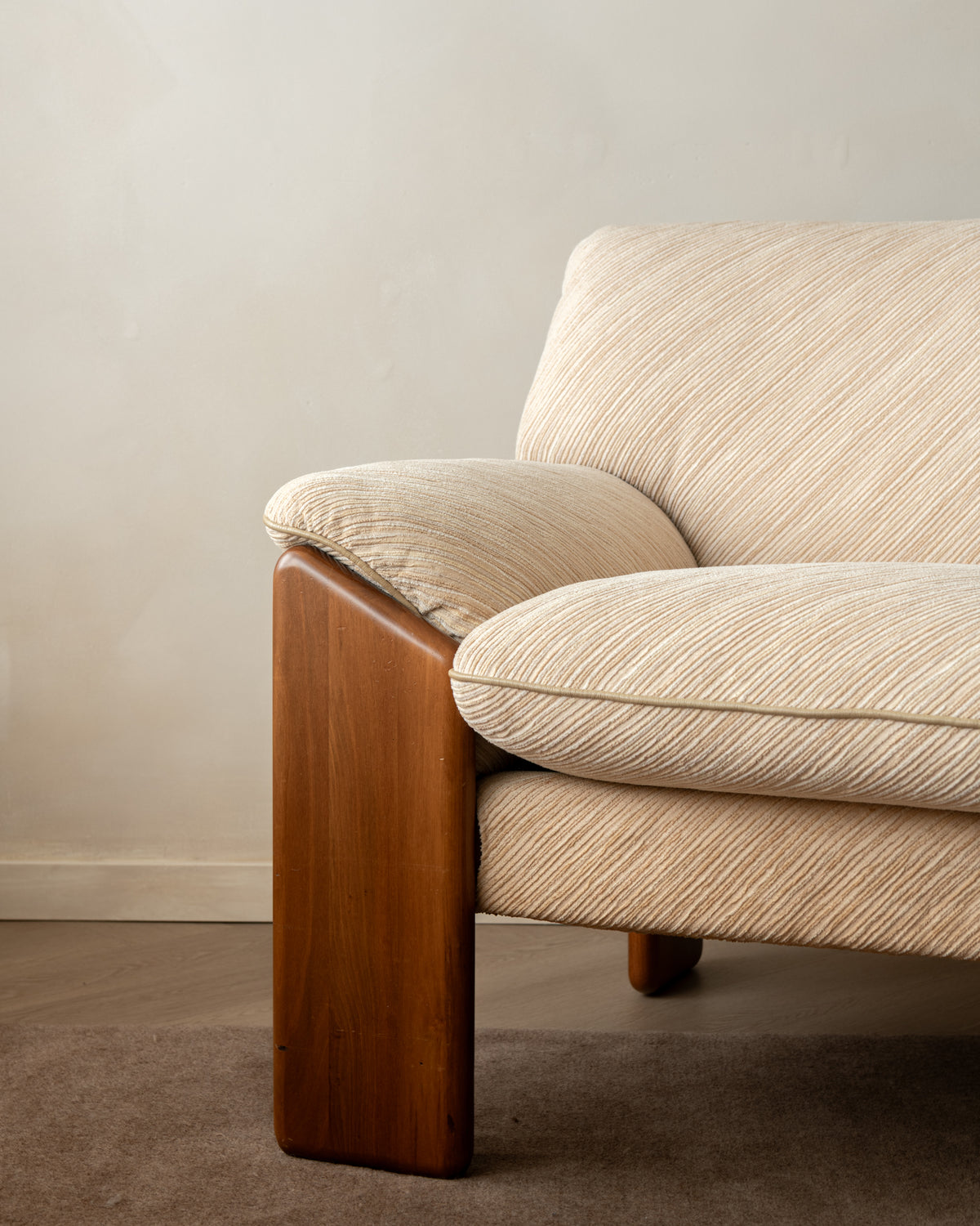 A close-up of a mid-century modern armchair featuring a warm brown wooden frame and textured beige fabric upholstery, set against a neutral beige wall and brown carpet.