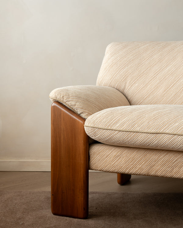 A close-up of a mid-century modern armchair featuring a warm brown wooden frame and textured beige fabric upholstery, set against a neutral beige wall and brown carpet.