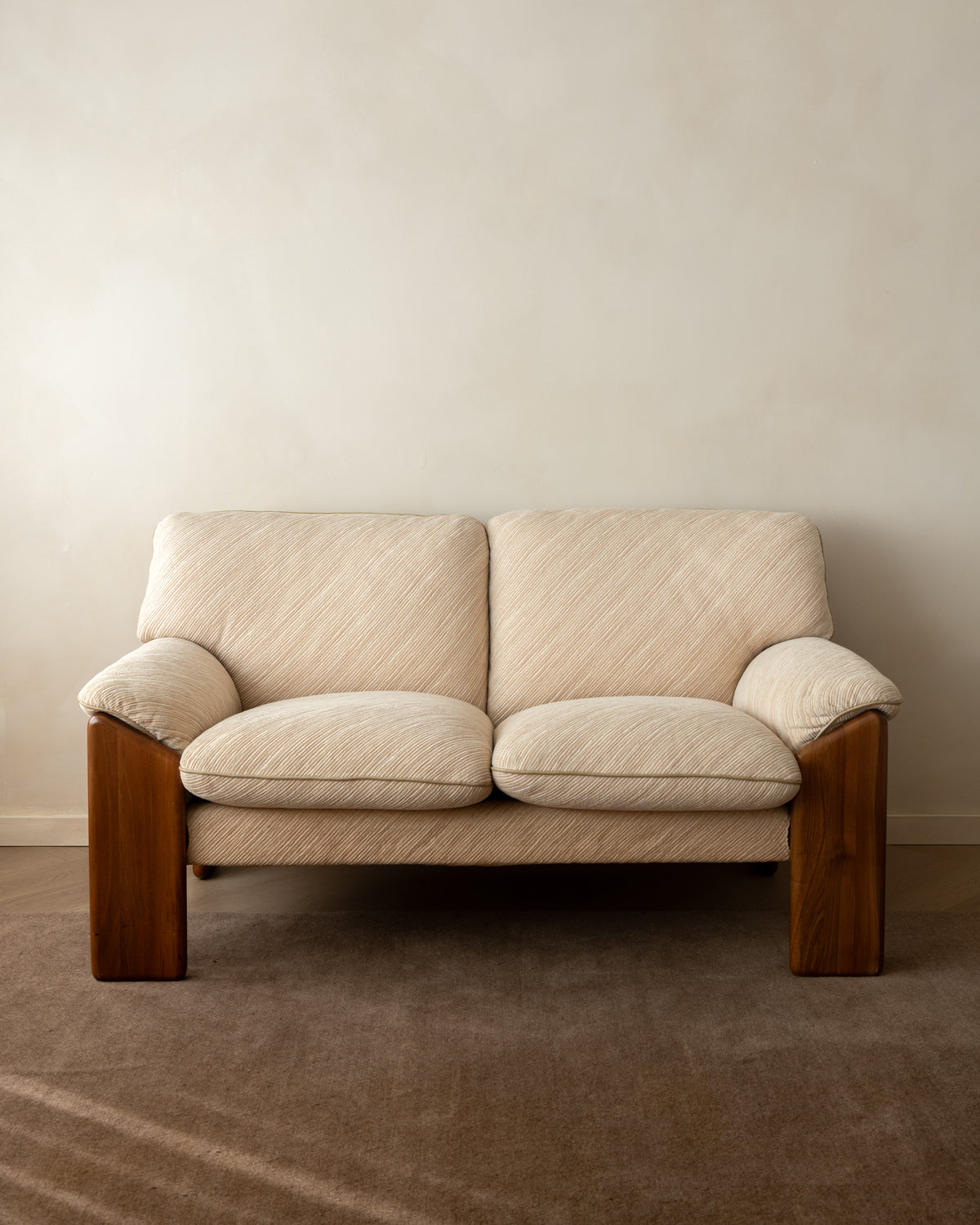 A two-seater loveseat sofa with cream-colored textured fabric upholstery and dark brown wooden legs, positioned against a neutral beige wall and brown carpet.
