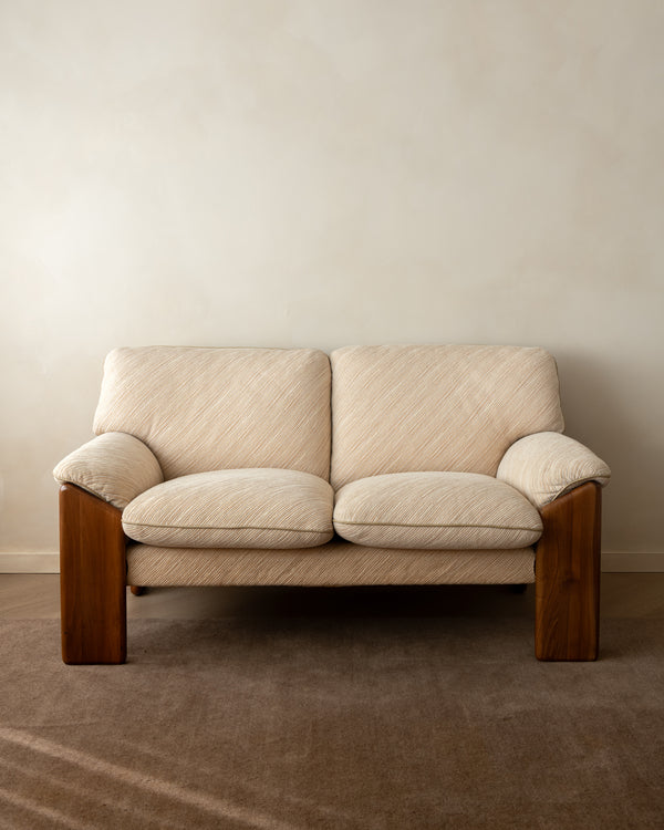 A two-seater loveseat sofa with cream-colored textured fabric upholstery and dark brown wooden legs, positioned against a neutral beige wall and brown carpet.