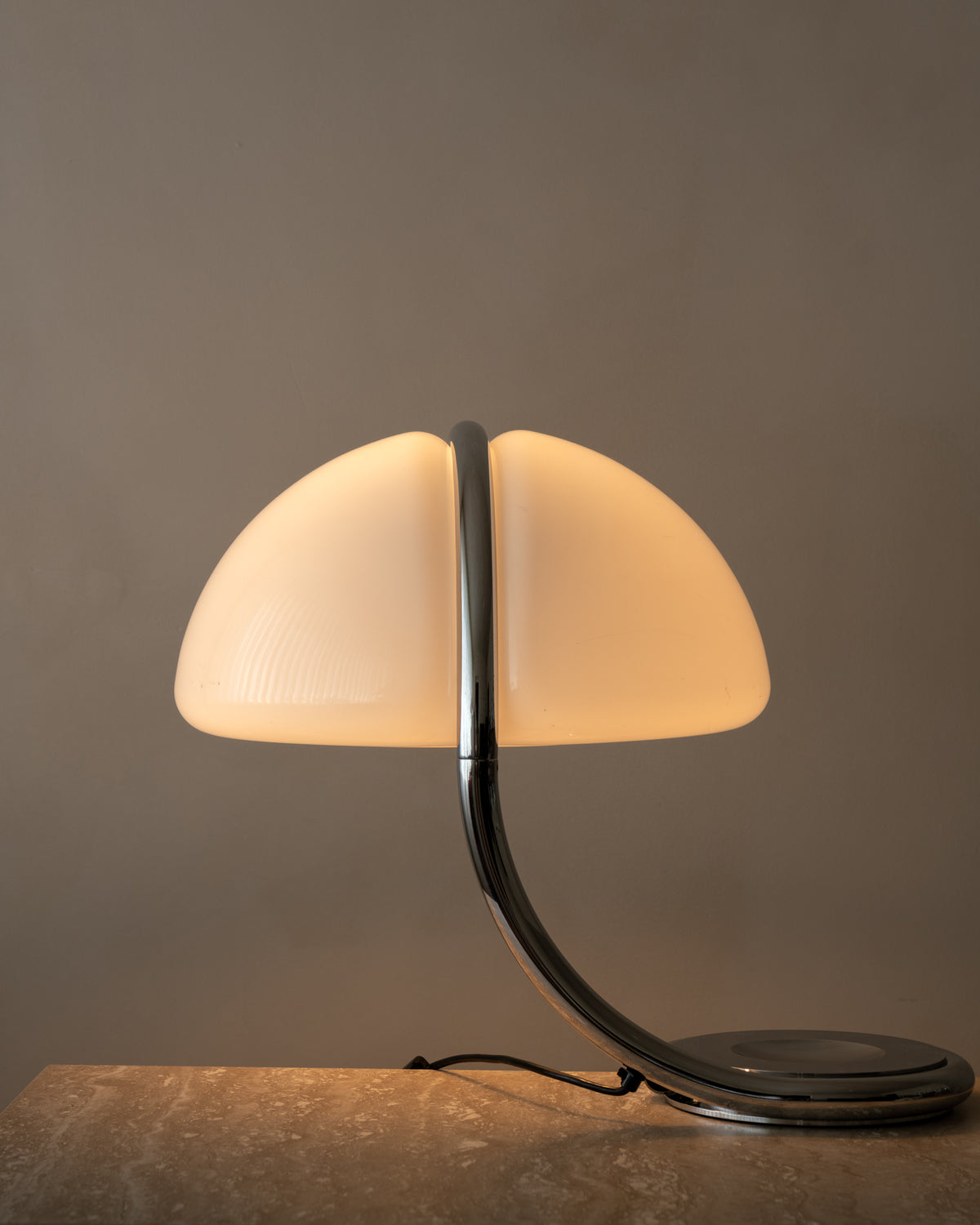A modern table lamp with a split dome-shaped frosted glass shade and a curved metallic base, shown lit against a warm gray background.