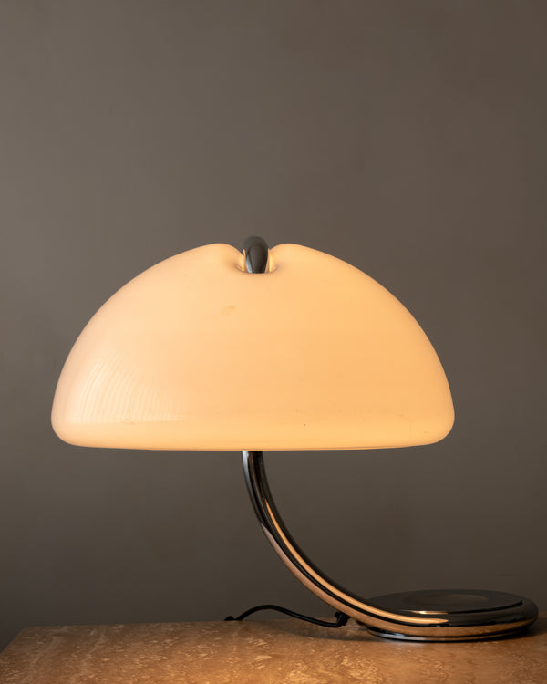 A vintage-style table lamp with a warm cream-colored glass dome shade and a curved black metal base, shown lit against a dark gray background.