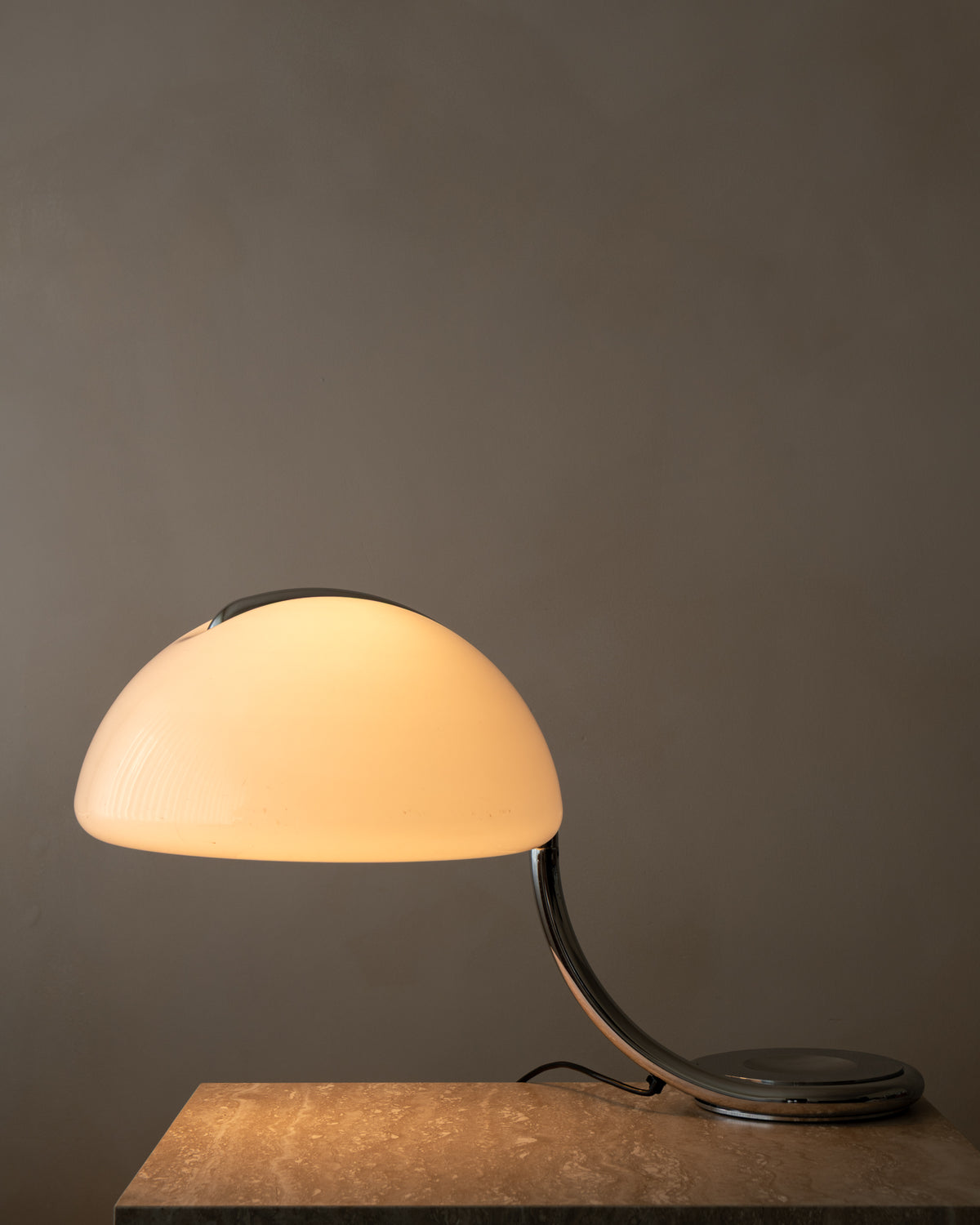A modern table lamp with a glowing warm white dome shade and a curved metallic base, shown resting on a wooden surface against a dark gray wall.