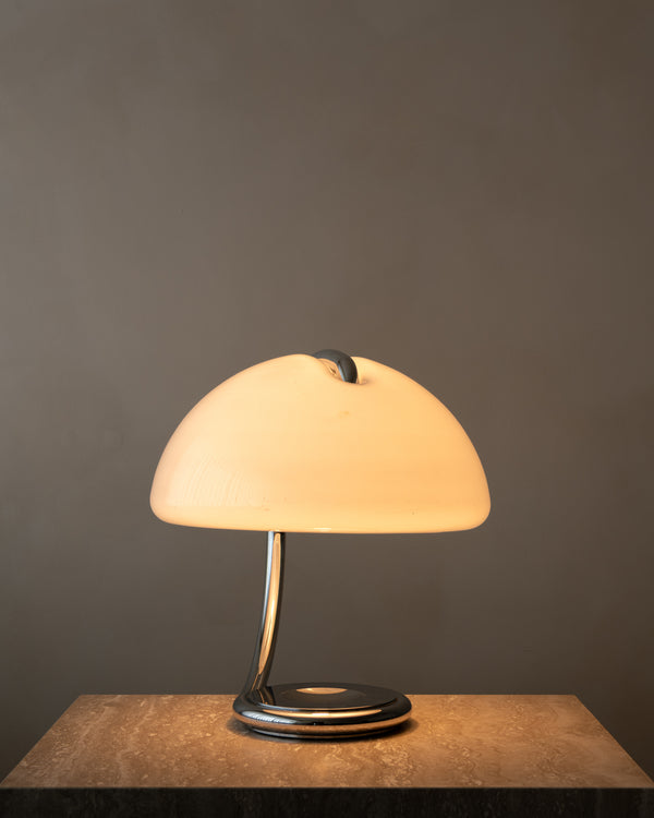 A modern Serpente table lamp with a glowing warm white dome shade and a polished chrome base, displayed on a textured stone surface against a dark gray background.