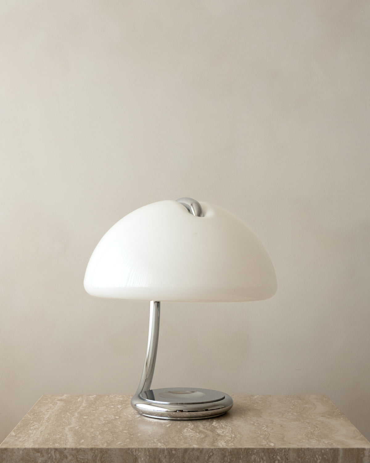 A modern Serpente table lamp with a glossy white dome shade and a polished chrome base, displayed on a textured stone surface against a neutral beige wall.