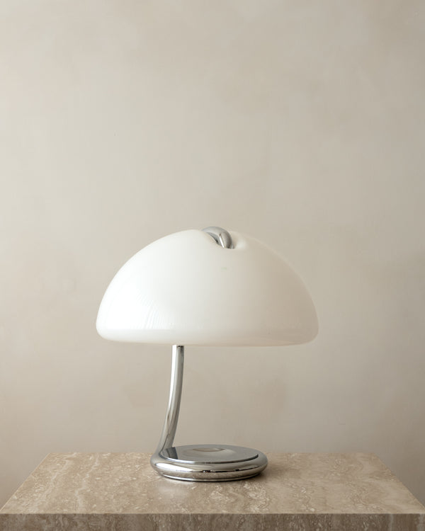 A modern Serpente table lamp with a glossy white dome shade and a polished chrome base, displayed on a textured stone surface against a neutral beige wall.