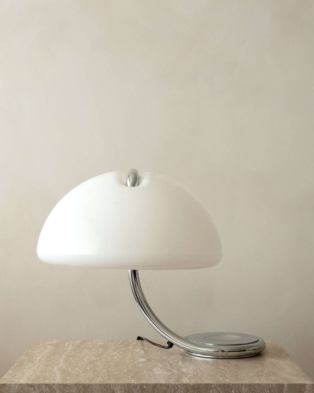 A mid-century modern table lamp with a glossy white dome shade and a curved chrome base, resting on a textured stone surface against a neutral beige wall.