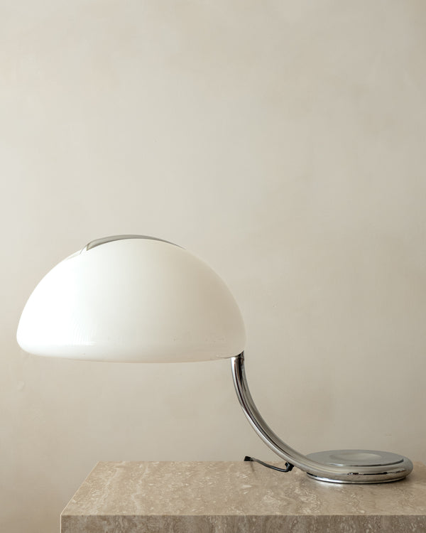 A modern table lamp with a glossy white dome-shaped shade and a curved chrome metal stem, resting on a light beige stone surface against a neutral beige wall.