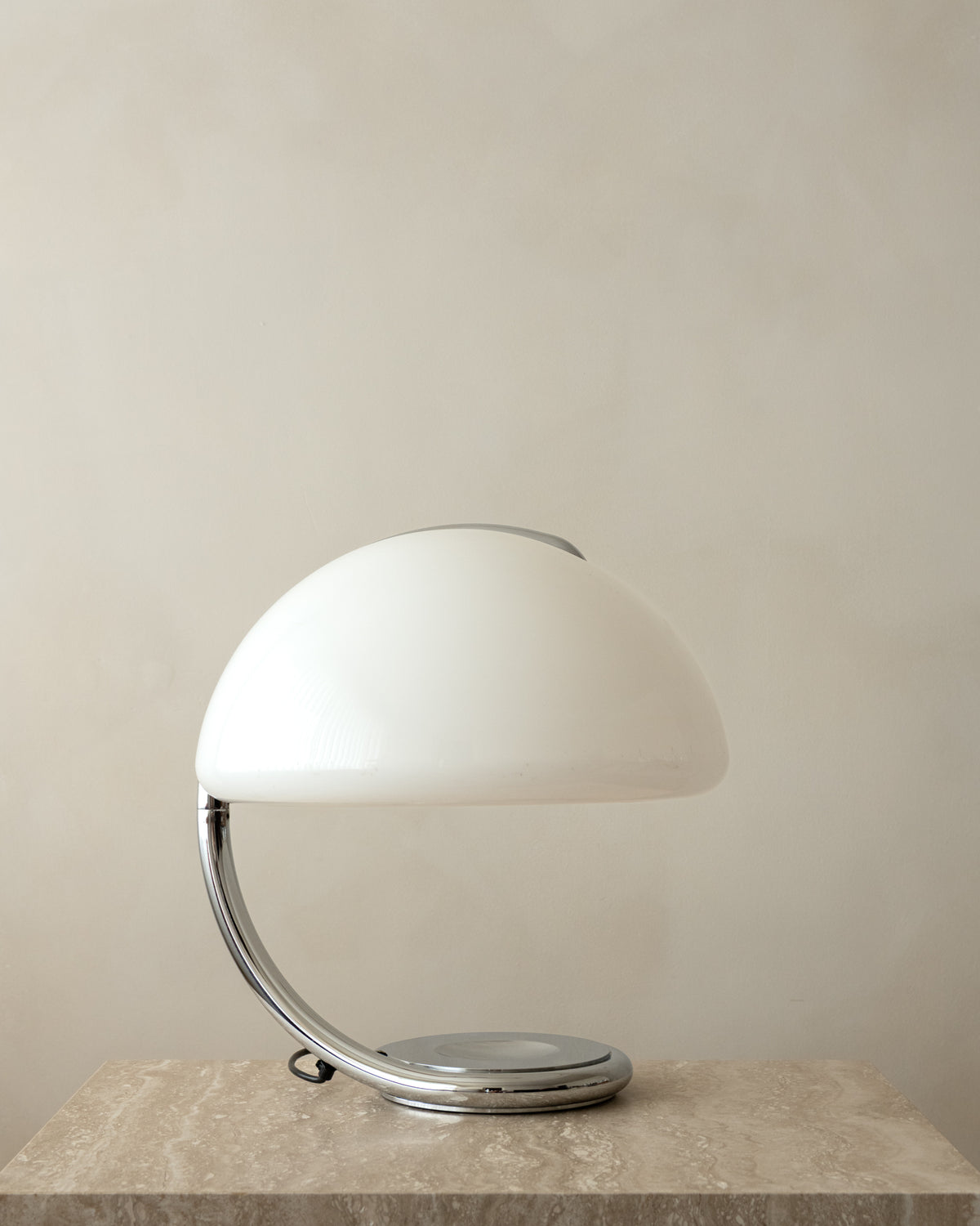 A modern table lamp with a glossy white dome-shaped shade and a curved chrome base, resting on a textured stone surface against a neutral beige wall.