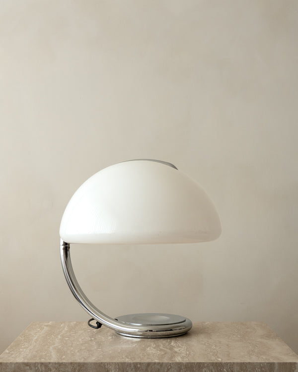 A modern table lamp with a glossy white dome-shaped shade and a curved chrome base, resting on a textured stone surface against a neutral beige wall.