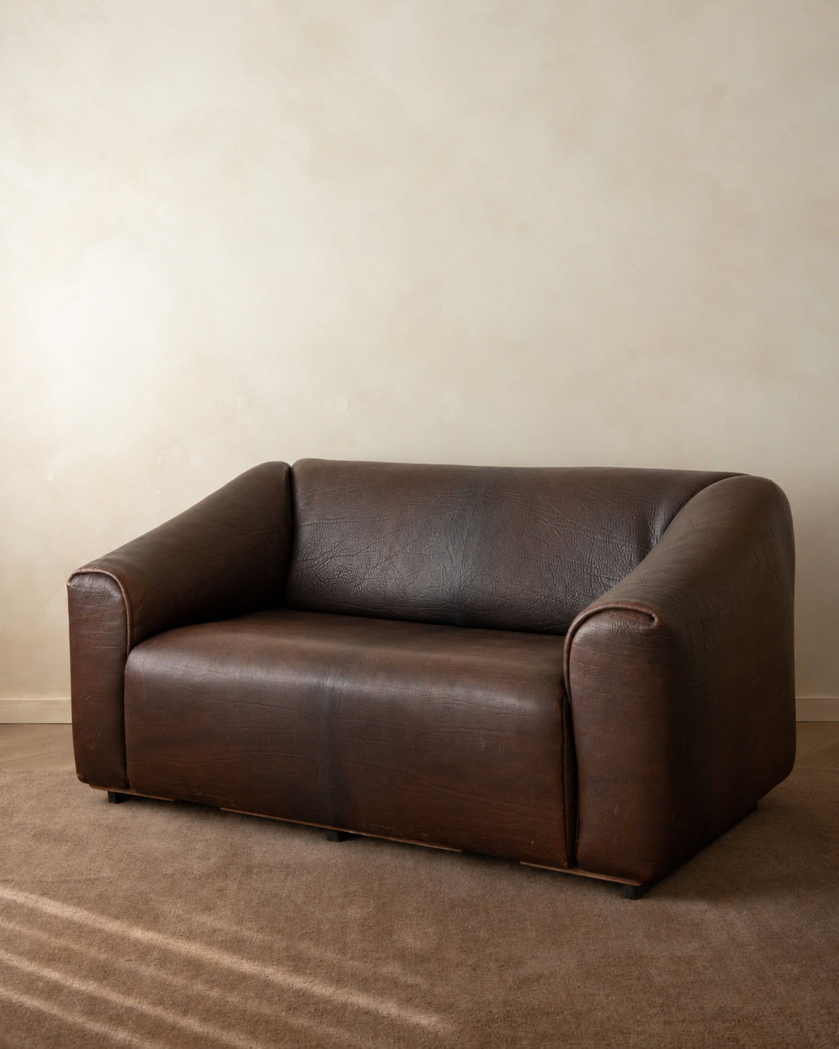 A dark brown leather loveseat sofa with rounded arms and a low back, positioned on a textured brown carpet against a neutral beige wall.