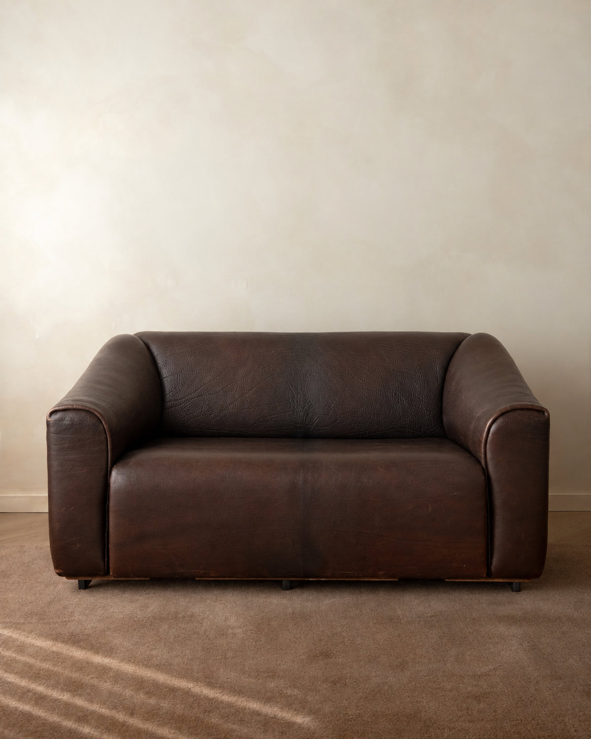A dark brown leather loveseat sofa placed against a textured beige wall on a brown carpeted floor.