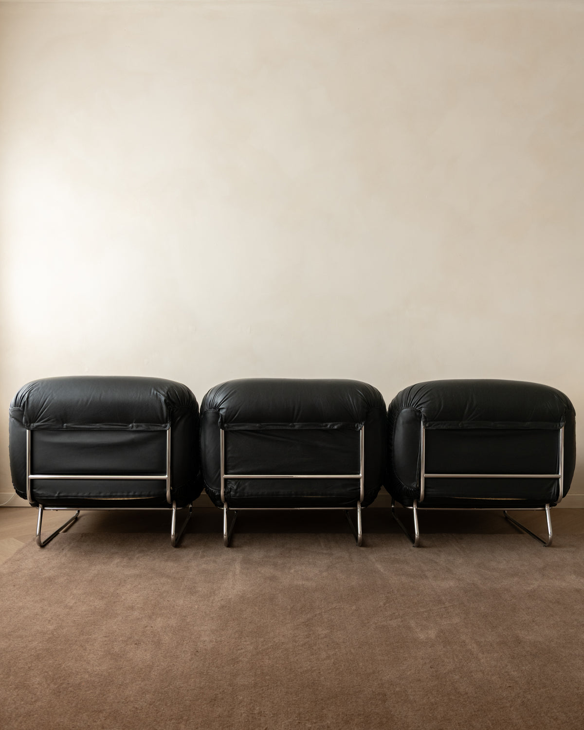 Three matching black leather lounge chairs with chrome legs arranged side-by-side against a warm beige wall and brown carpet.